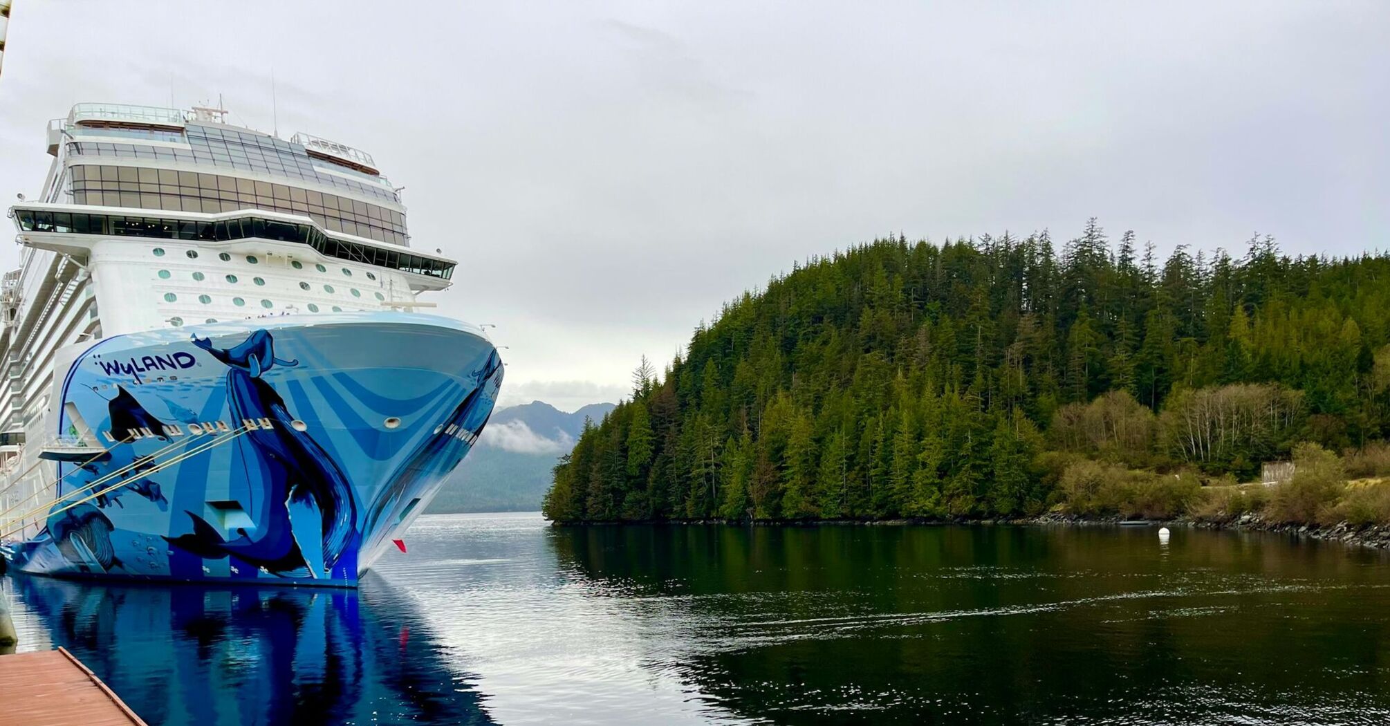 Norwegian Cruise Line ship docked near forested Alaskan coastline