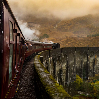 Steam train crossing a historic stone viaduct through misty hills