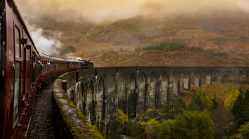 Steam train crossing a historic stone viaduct through misty hills