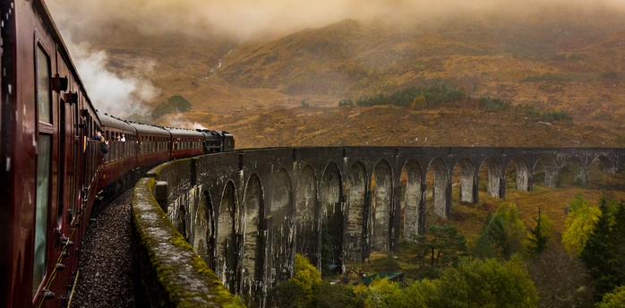 Steam train crossing a historic stone viaduct through misty hills
