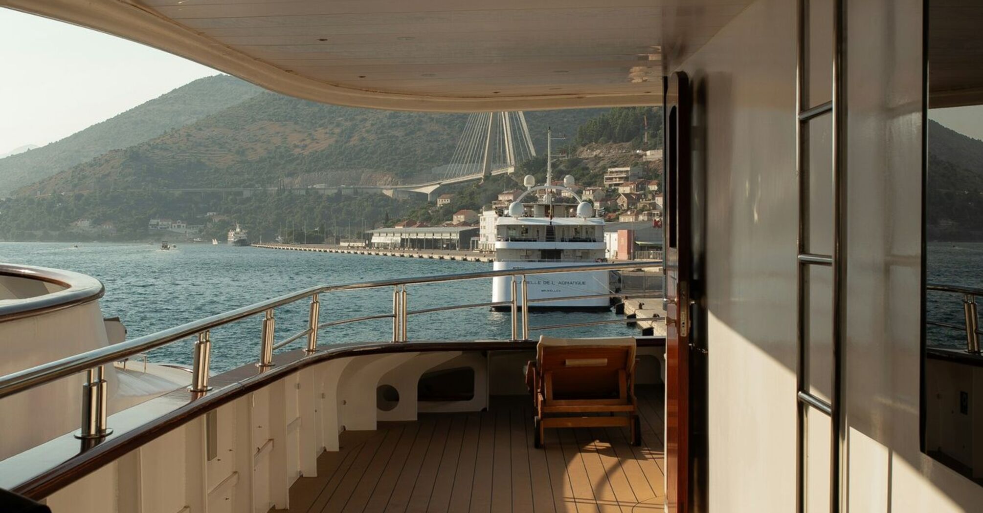 Empty deck of a cruise ship docked in a coastal European port with hills and a bridge in the background