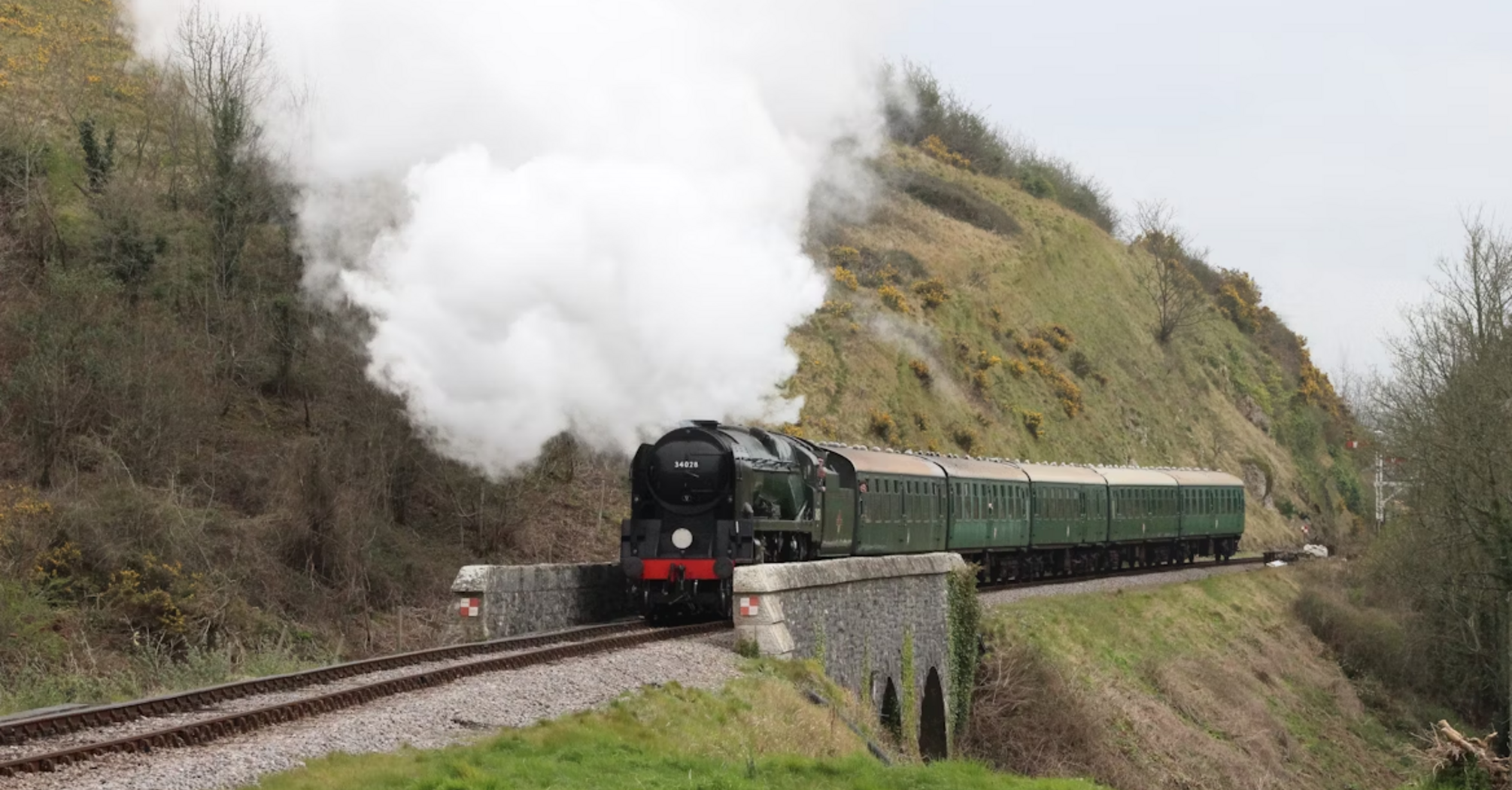 A steam train crossing a small stone bridge through a green countryside valley, releasing white smoke