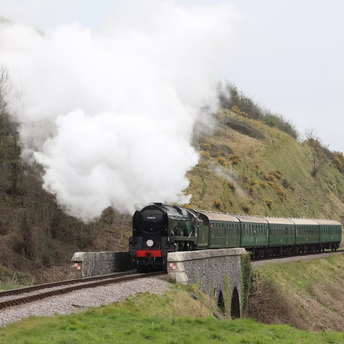 A steam train crossing a small stone bridge through a green countryside valley, releasing white smoke