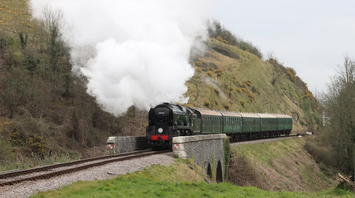 A steam train crossing a small stone bridge through a green countryside valley, releasing white smoke