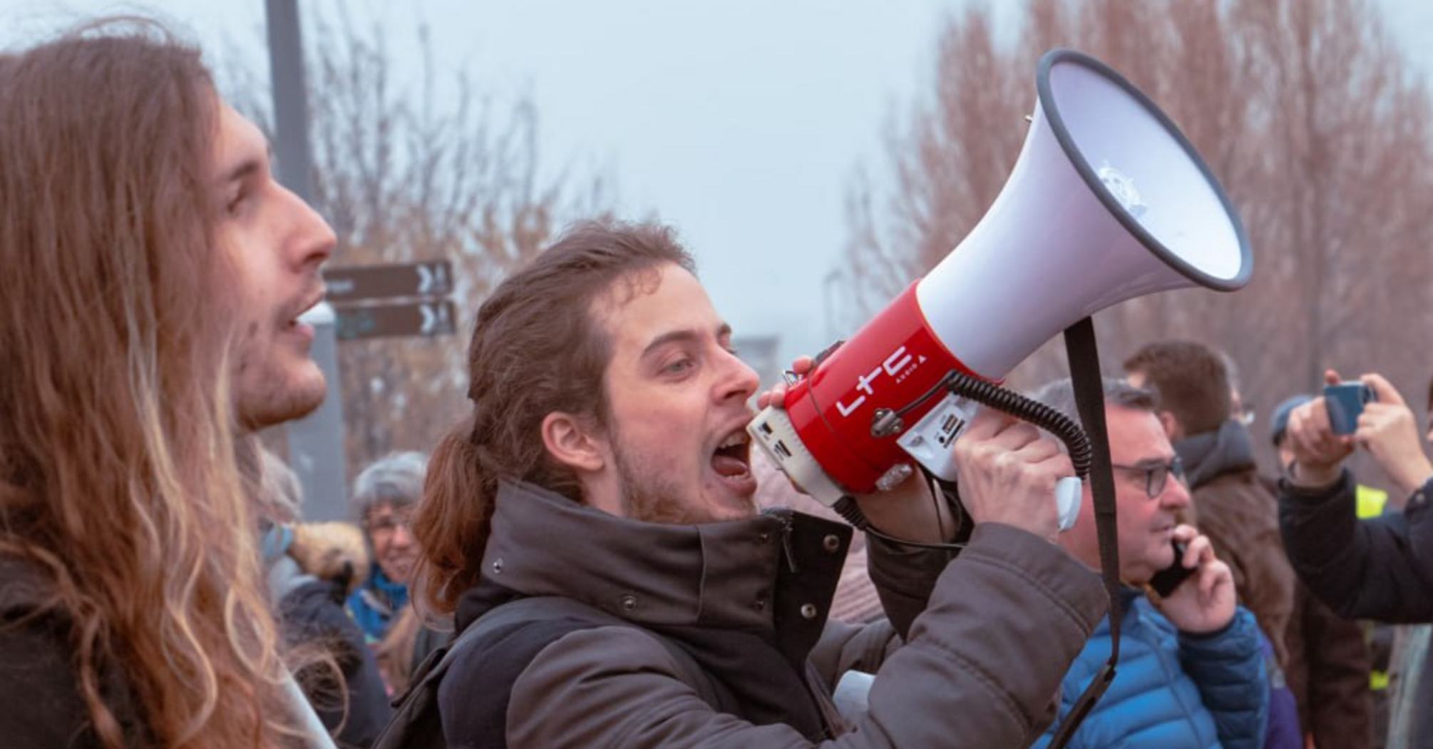 A man shouting into a megaphone during a protest with a crowd in the background