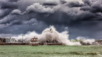 Waves crashing over a harbor wall during a severe storm under dark clouds