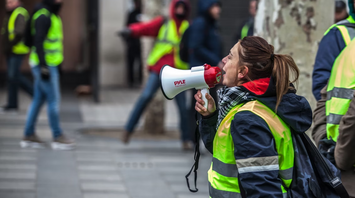 A woman in a yellow vest shouting into a megaphone during a street protest