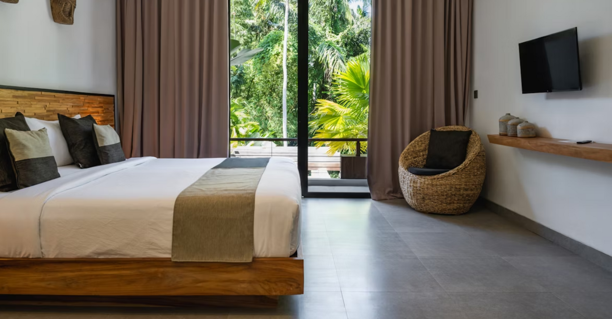Modern tropical hotel room with a large bed, woven chair, and a view of lush greenery through floor-to-ceiling glass doors