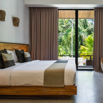 Modern tropical hotel room with a large bed, woven chair, and a view of lush greenery through floor-to-ceiling glass doors