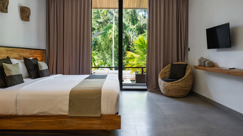 Modern tropical hotel room with a large bed, woven chair, and a view of lush greenery through floor-to-ceiling glass doors
