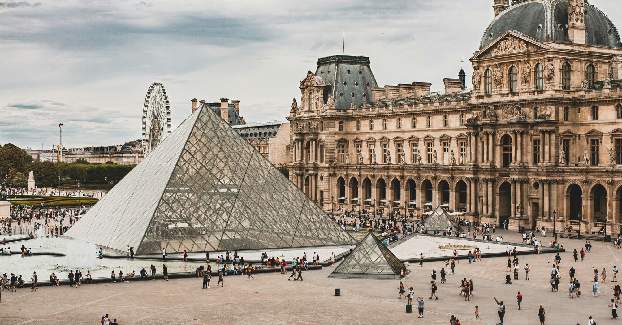 A wide view of the Louvre pyramid complex with visitors walking around the courtyard in Paris