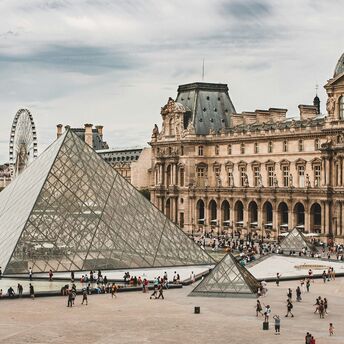 A wide view of the Louvre pyramid complex with visitors walking around the courtyard in Paris