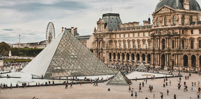 A wide view of the Louvre pyramid complex with visitors walking around the courtyard in Paris
