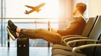 Traveller sitting at an airport gate with legs on luggage, watching a plane take off