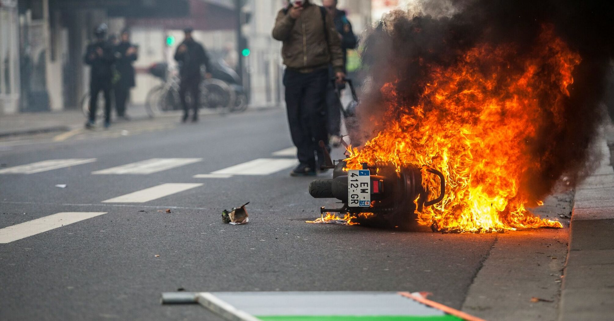 Motorcycle burning in city street during unrest with smoke and pedestrians nearby