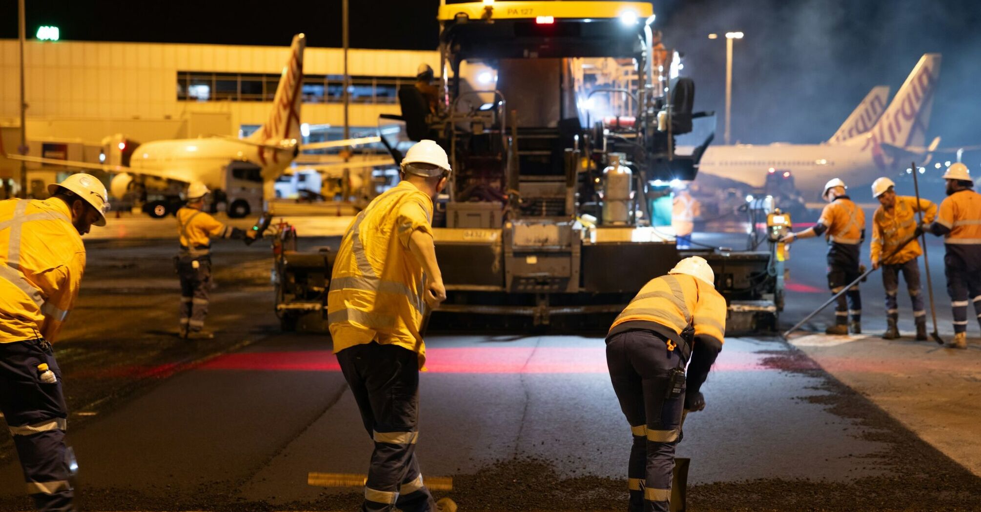 Airport workers resurfacing runway at night near terminal