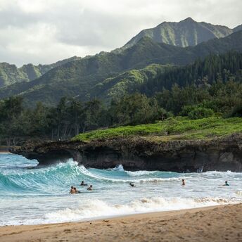 Surfers in ocean waves with green mountains behind on a Hawaii beach