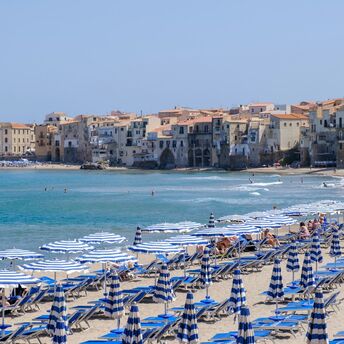 Cefalù beach with striped umbrellas and historic seaside town in Italy